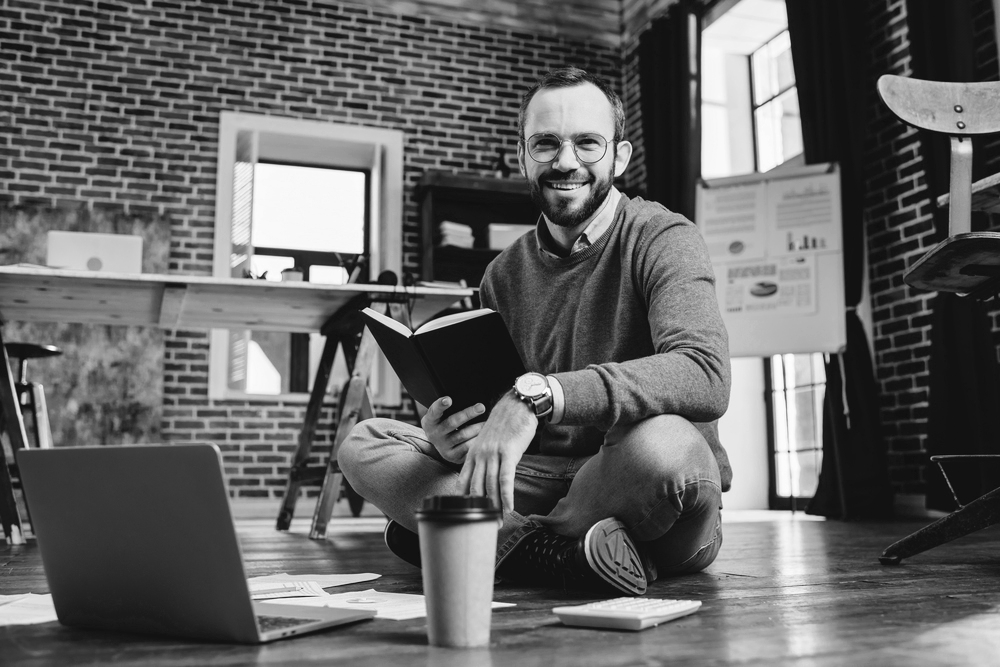 Business owner reading a book on the floor infront of a laptop with a coffee